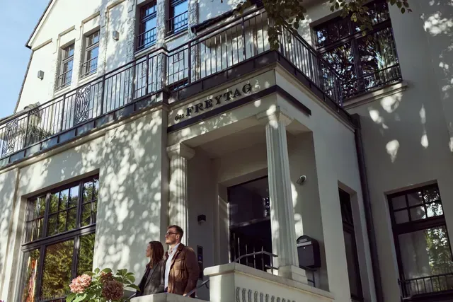 Ein Mann und eine Frau stehen auf einem Balkon vor einem großen weißen Gebäude.