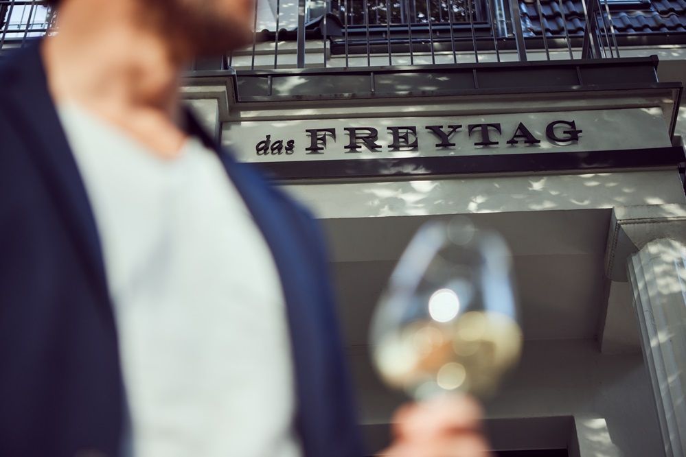 Ein Mann hält ein Glas Wein vor einem Schild mit der Aufschrift „Das Freytag“