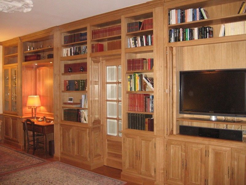 Un mur d'étagères en bois encastrées contenant des livres, un petit coin bureau avec une lampe et une grande télévision.