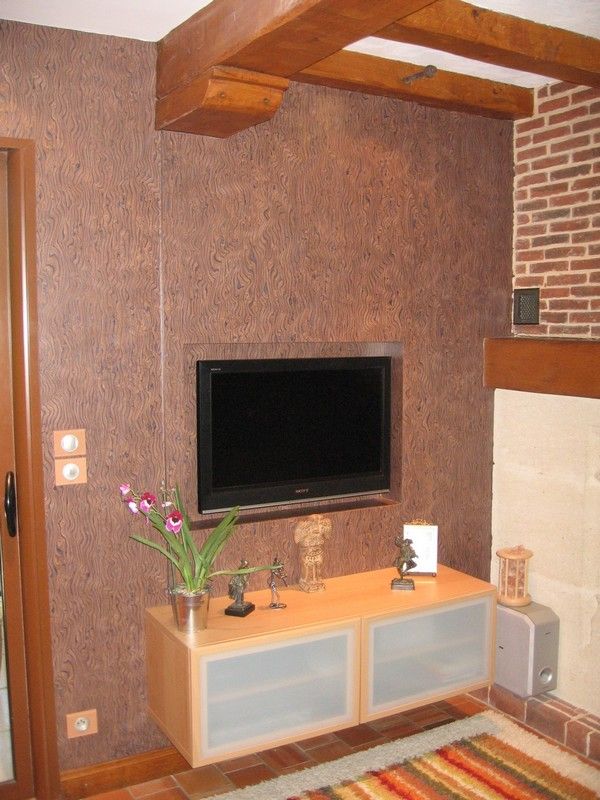 Dans une pièce, un téléviseur fixé au mur est placé au-dessus d'une console en bois clair ornée d'objets décoratifs, le tout adossé à un mur texturé brun.