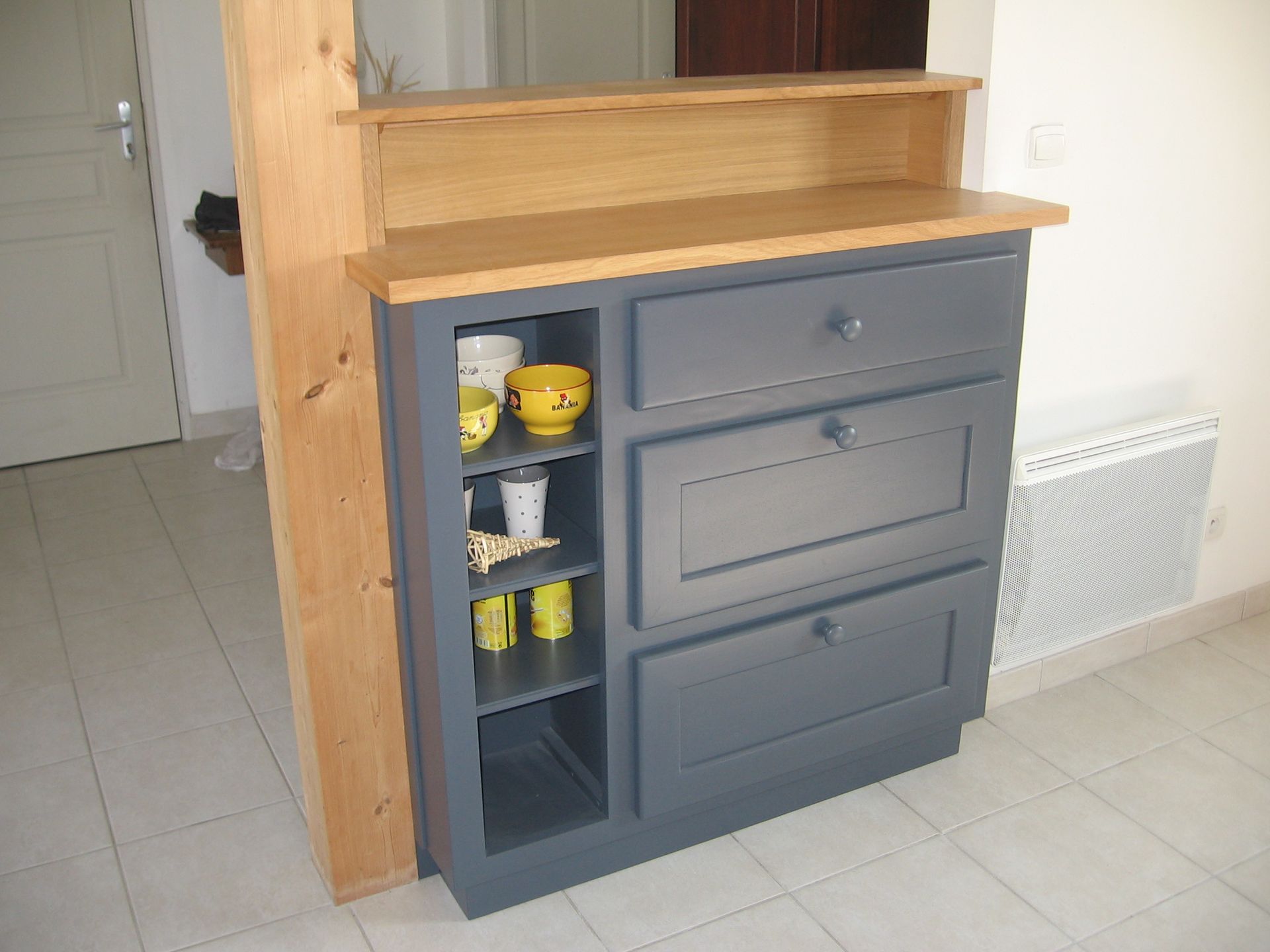 Un meuble gris avec une étagère latérale ouverte et trois tiroirs, surmonté d'un comptoir en bois, dans un intérieur de maison.