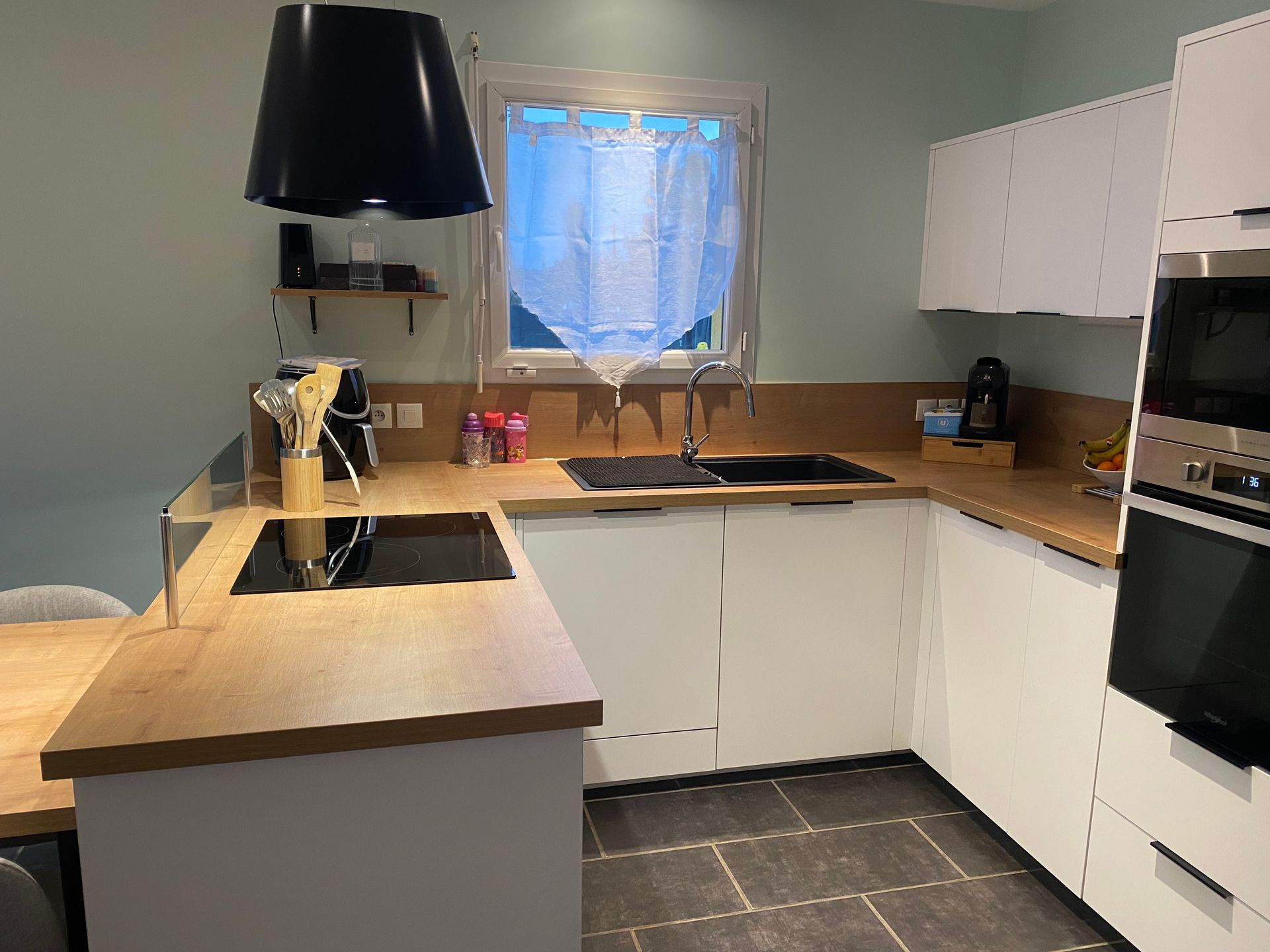 Cuisine moderne avec comptoirs en bois clair, placards blancs, sols en carrelage de pierre foncée, hotte et appareils électroménagers noirs.