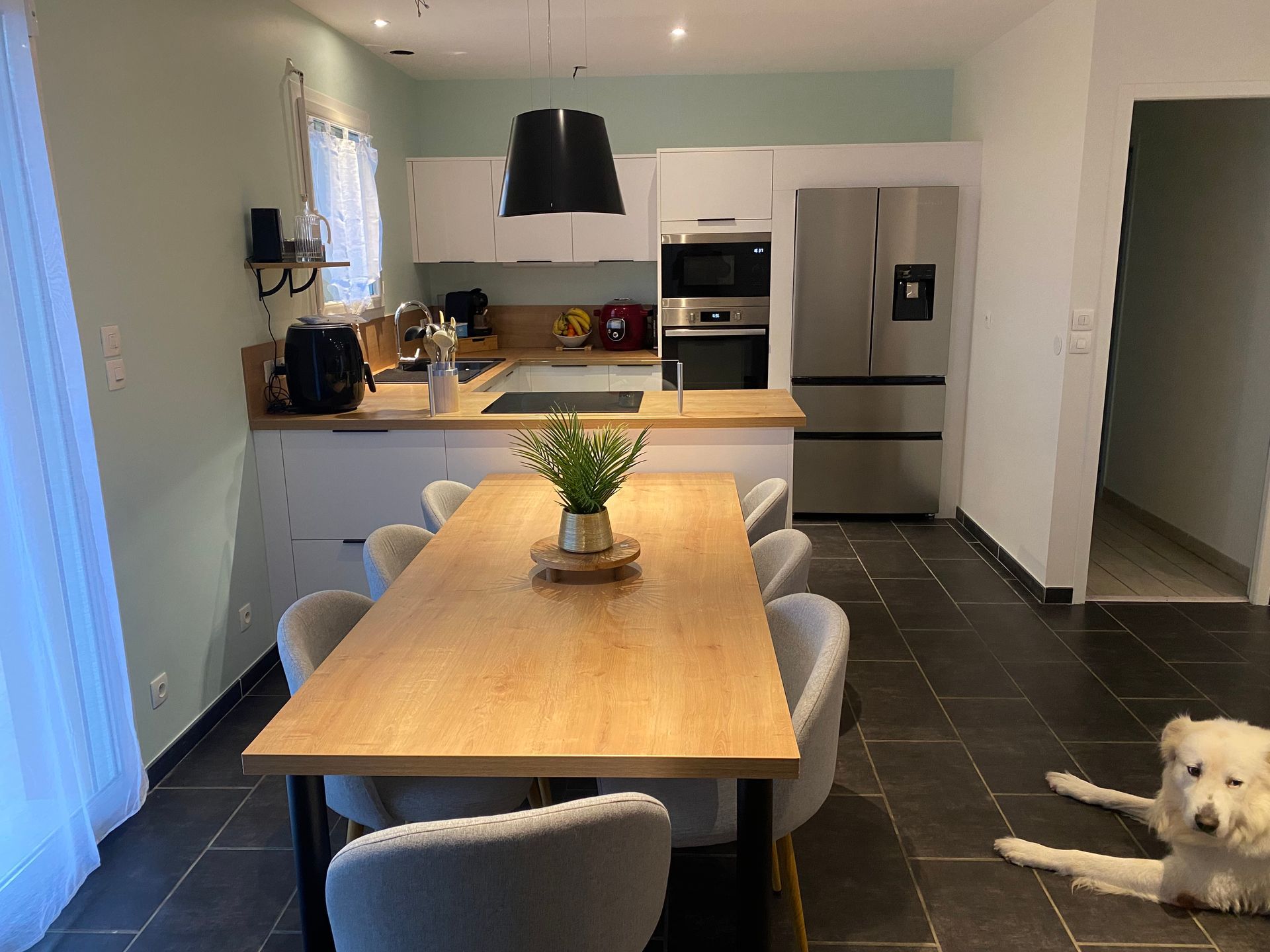 Une cuisine moderne avec une table à manger en bois, des chaises gris clair et un chien blanc allongé sur le sol carrelé foncé.