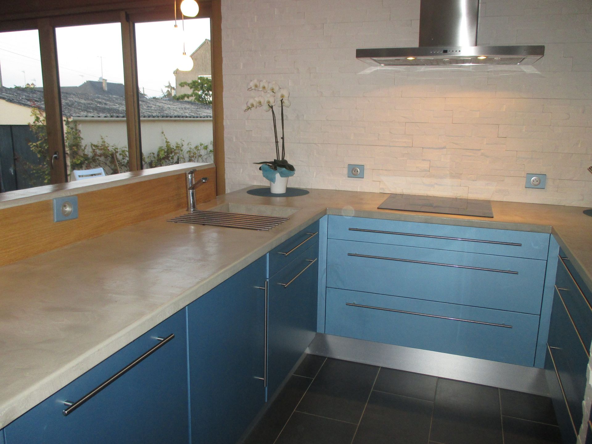 Une cuisine en forme de U avec des placards bleus, un comptoir beige, une hotte en acier inoxydable et des carreaux de sol noirs.