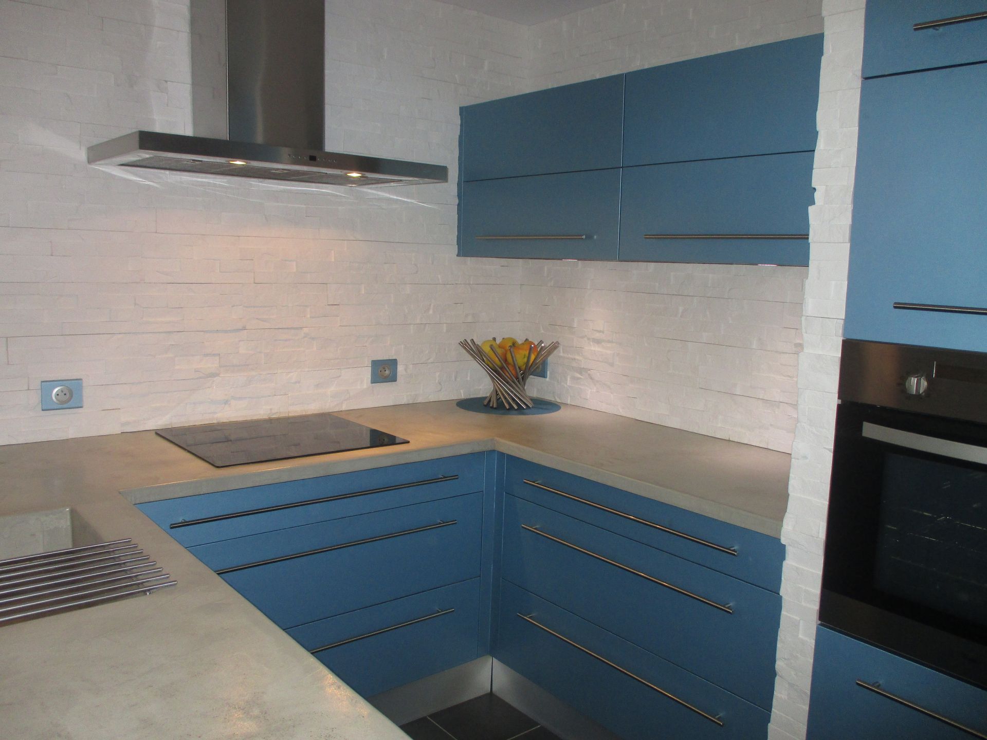 Cuisine moderne en forme de U avec placards bleus, comptoirs en béton, murs blancs texturés et hotte en acier inoxydable.