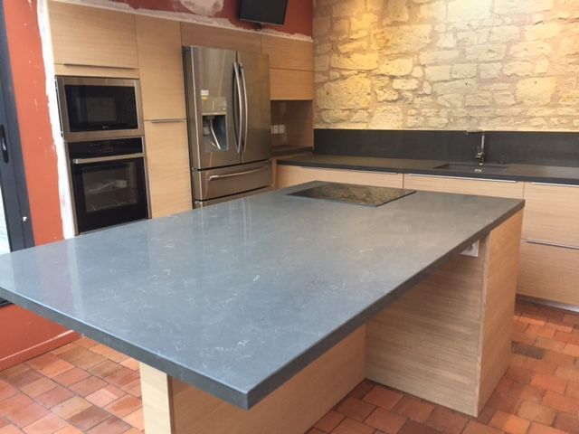 Cuisine moderne avec un grand îlot gris, des armoires en bois clair, un réfrigérateur en acier inoxydable et un mur en pierre.