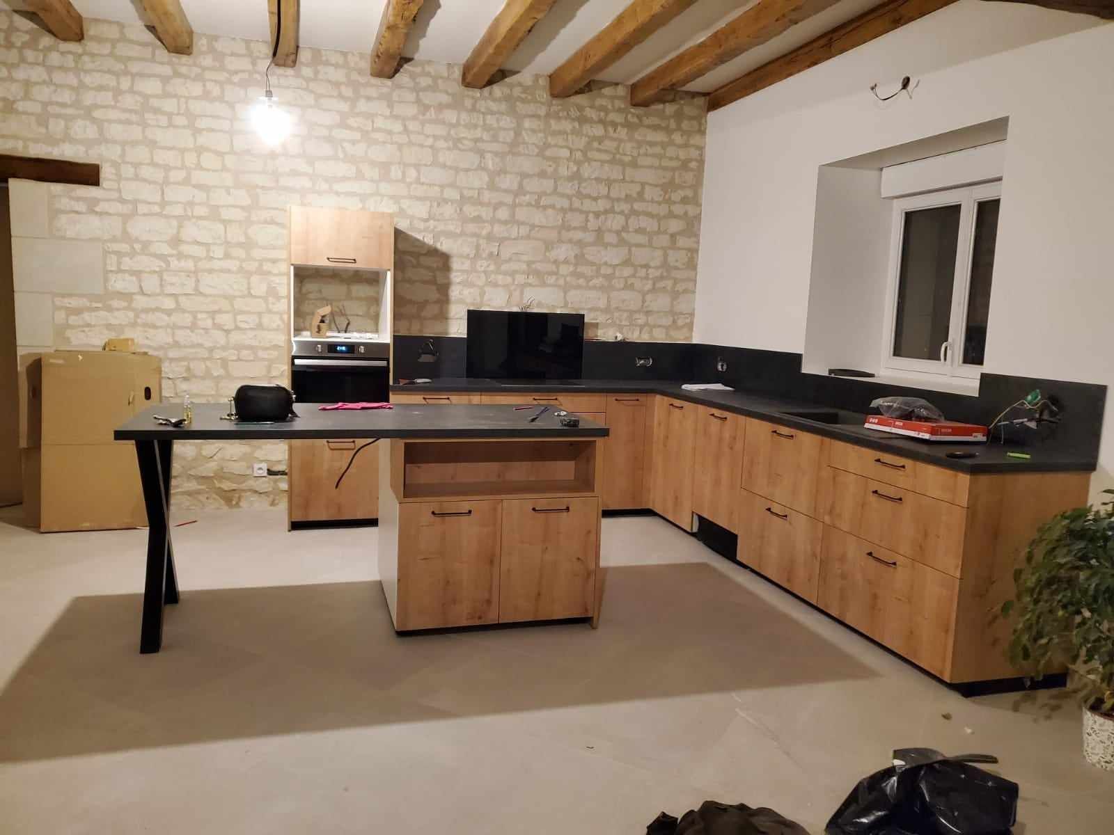 Une cuisine moderne aux tons boisés, avec un grand îlot central et des plans de travail foncés, le tout adossé à un mur en pierre rustique.