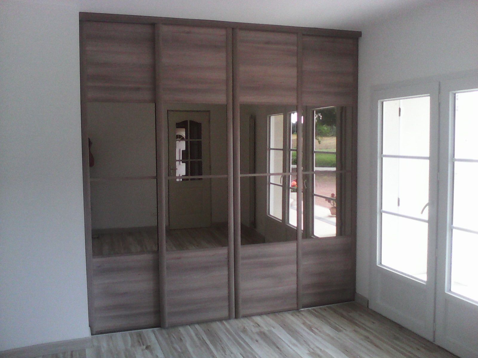 Une grande armoire coulissante à quatre panneaux, avec une finition imitation bois et des sections en miroir, intégrée dans une pièce aux tons clairs.