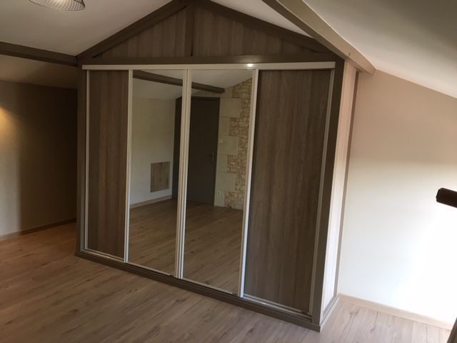 Une armoire moderne avec des portes en imitation bois et un miroir central, intégrée dans une pièce mansardée au plafond en pente.