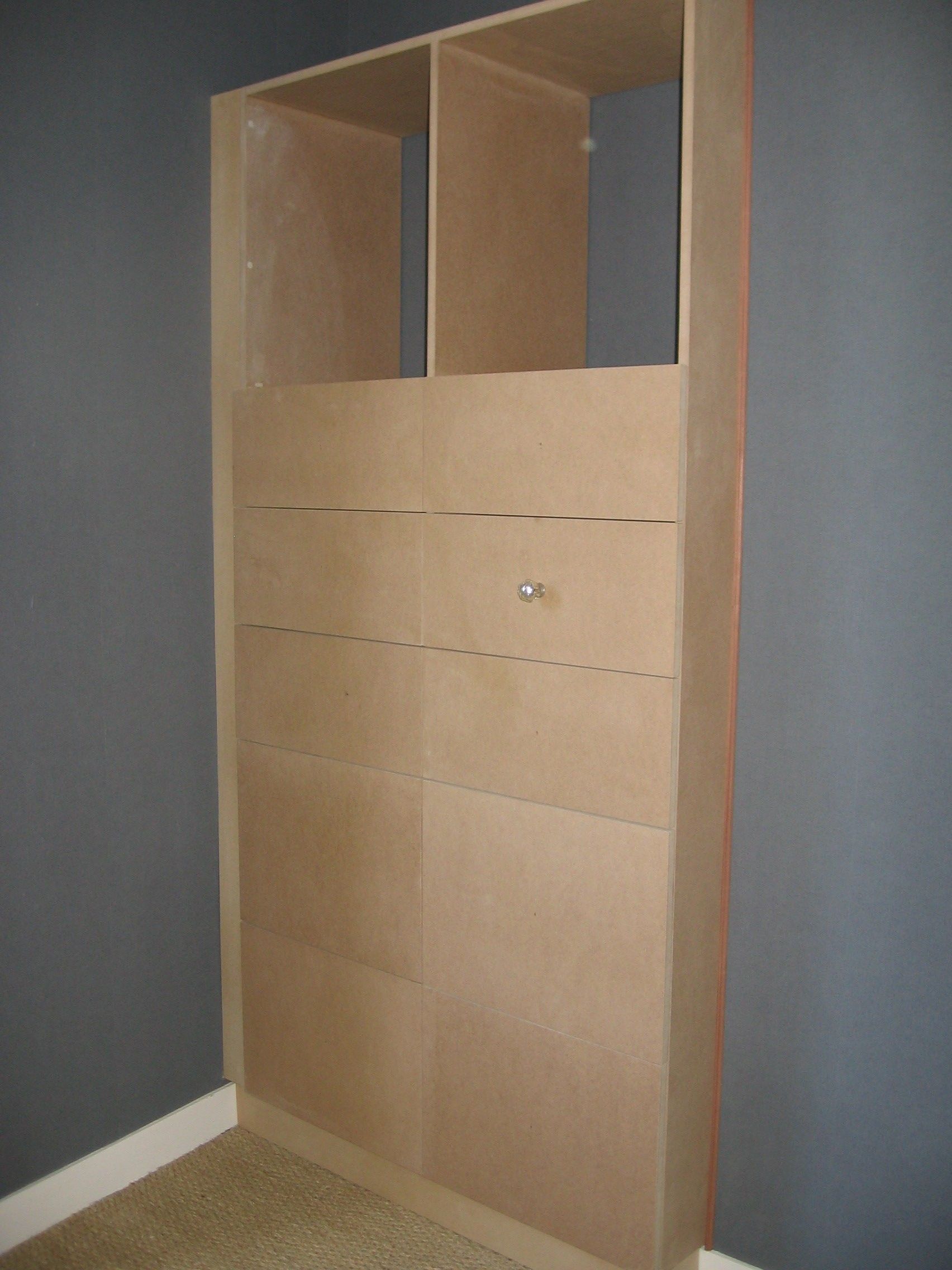 Meuble de rangement en bois brut contre un mur gris, avec une étagère supérieure ouverte et une partie inférieure munie d'une seule poignée.