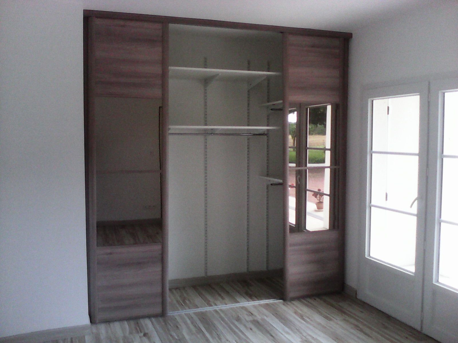 Un meuble de rangement à ossature bois avec étagères ouvertes et espace penderie central, placé contre un mur à côté de portes vitrées.