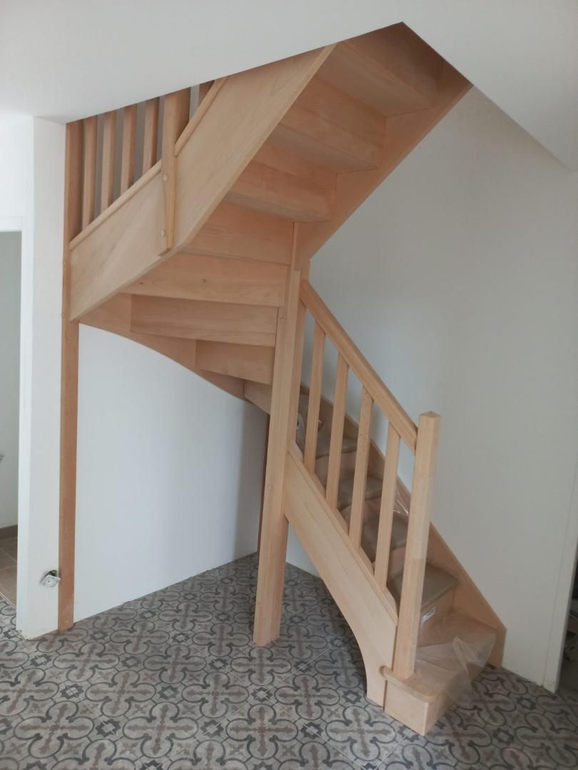 Un escalier quart tournant en bois clair, doté d'une rampe simple, se dresse devant des murs blancs sur un sol carrelé à motifs.