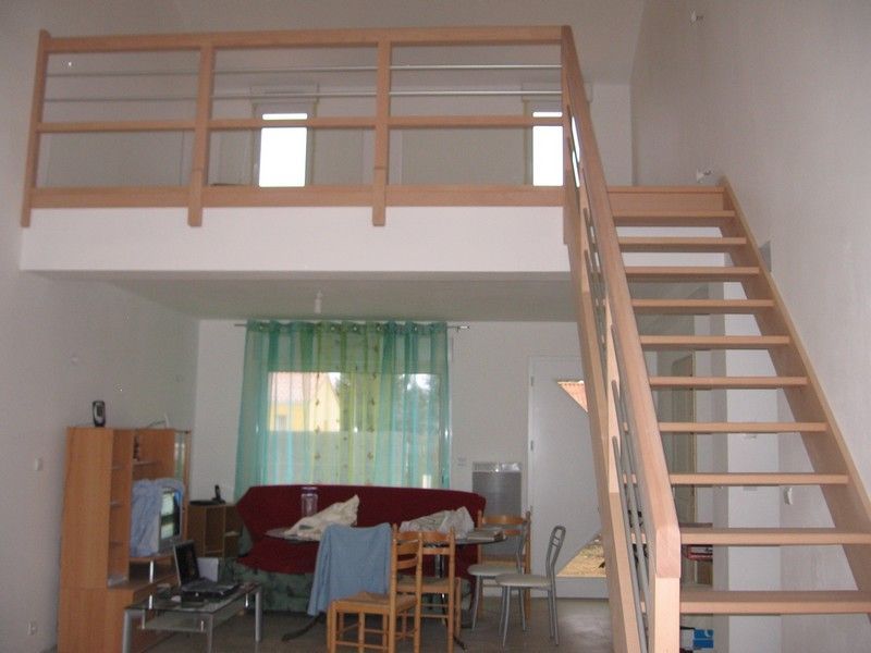 Une mezzanine avec une balustrade en bois surplombe un salon avec un canapé rouge, une table en bois et un escalier menant à l'étage.