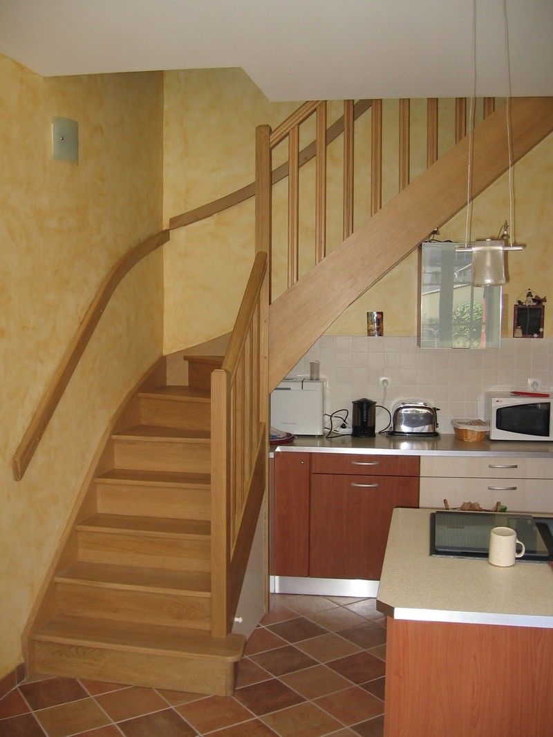 Un escalier en bois clair mène à l'étage depuis une cuisine équipée de placards en bois et d'un sol carrelé.