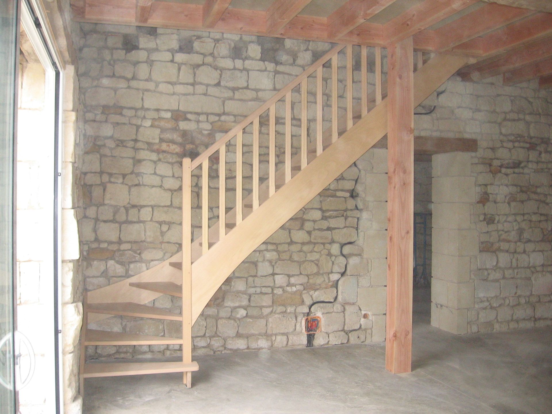 Un escalier en bois avec une simple rampe se dresse dans une pièce inachevée aux murs de pierre apparente et aux poutres de plafond en bois.