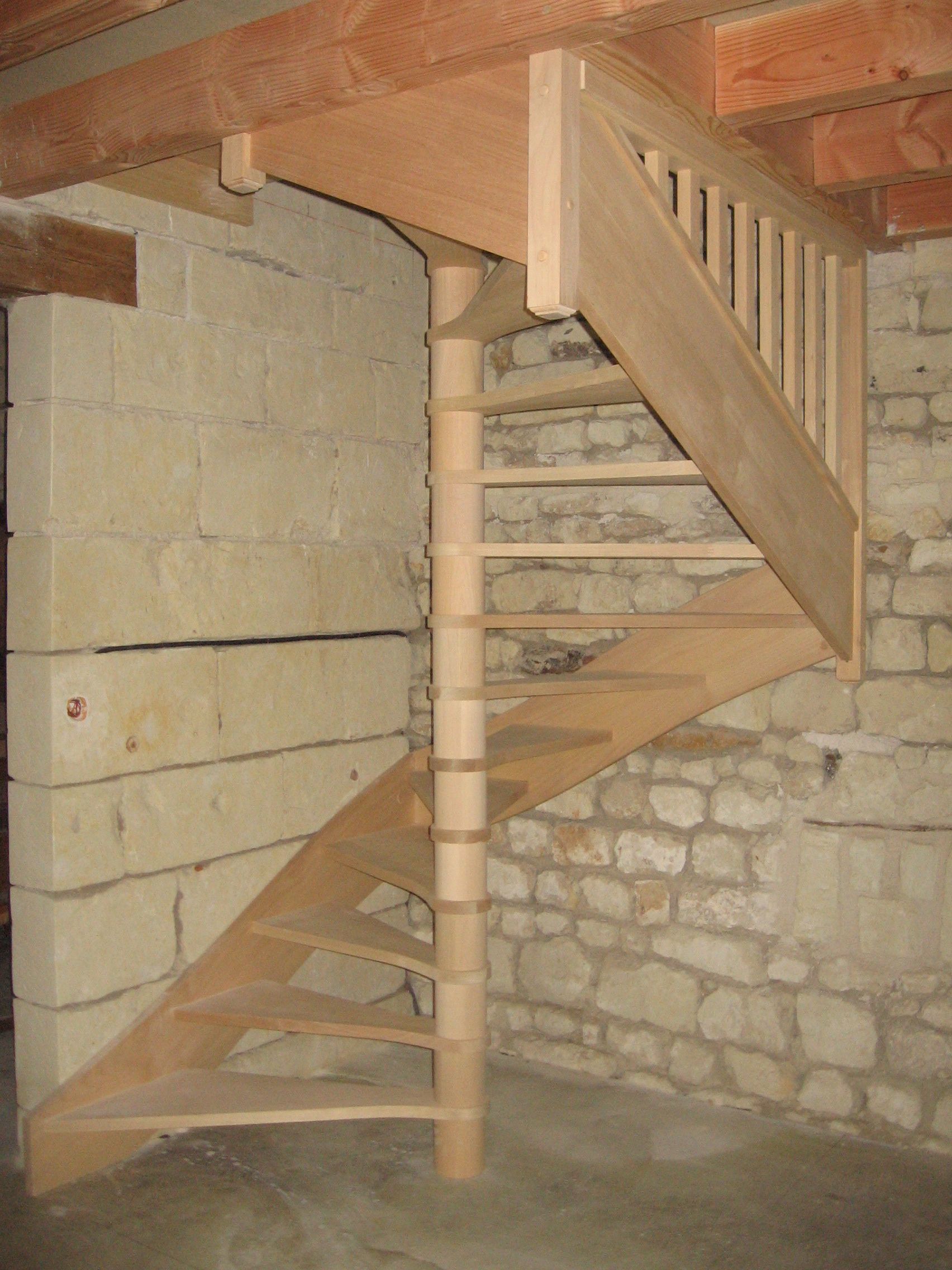 Un escalier en colimaçon en bois, inachevé, se dresse contre un mur de pierre brute dans un espace intérieur sous des poutres apparentes au plafond.