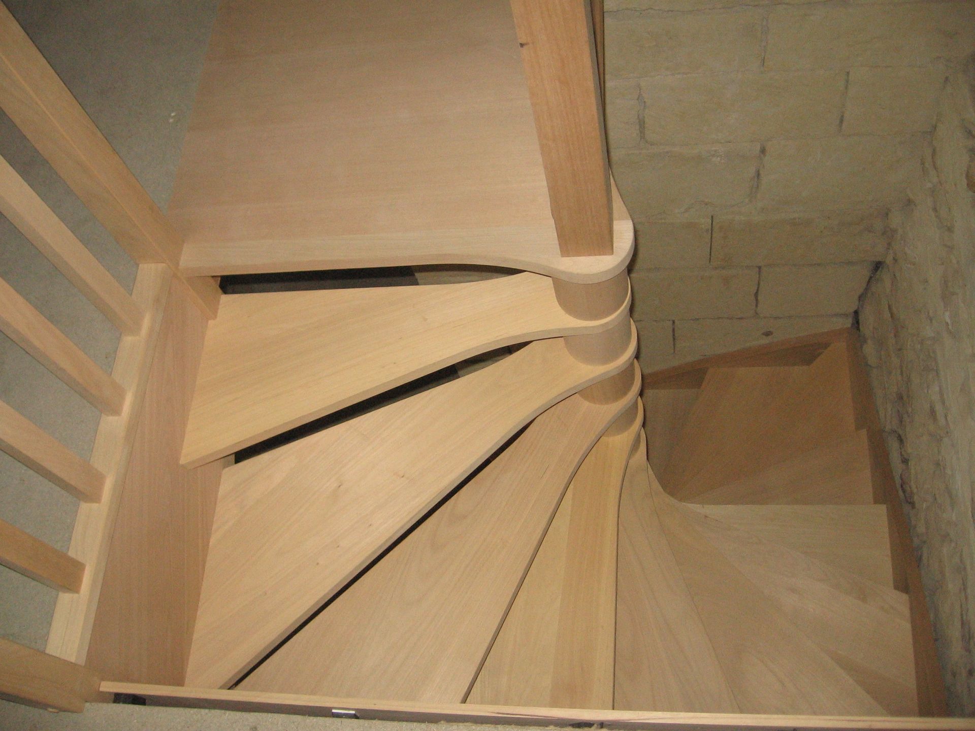 Vue en plongée d'un escalier en colimaçon en bois clair adossé à un mur de pierre, caractérisé par des marches étroites en forme de coin.