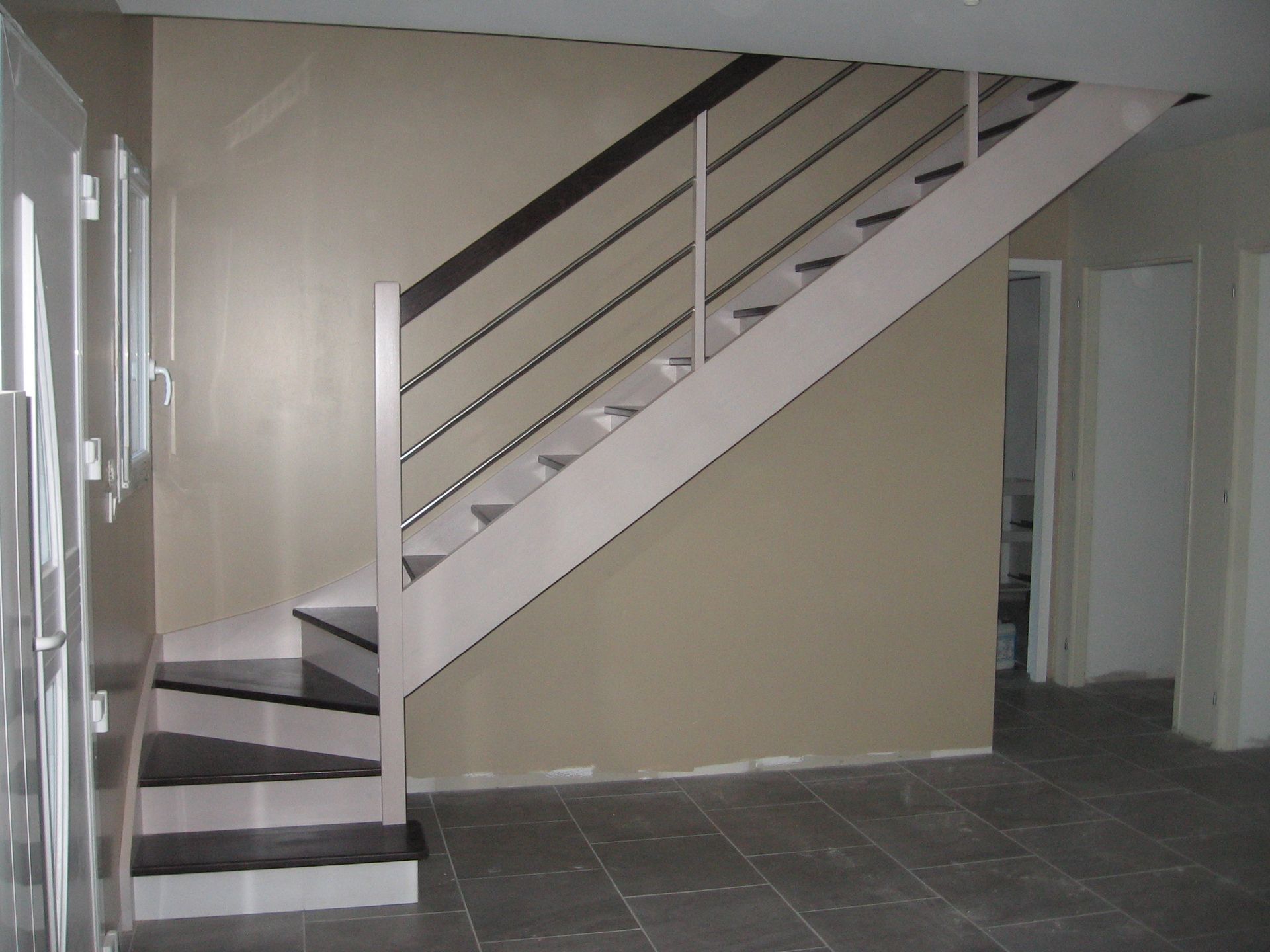 Un escalier moderne avec des marches en bois foncé, des supports blancs et une rampe en métal dans une pièce aux murs beiges et aux carreaux gris.