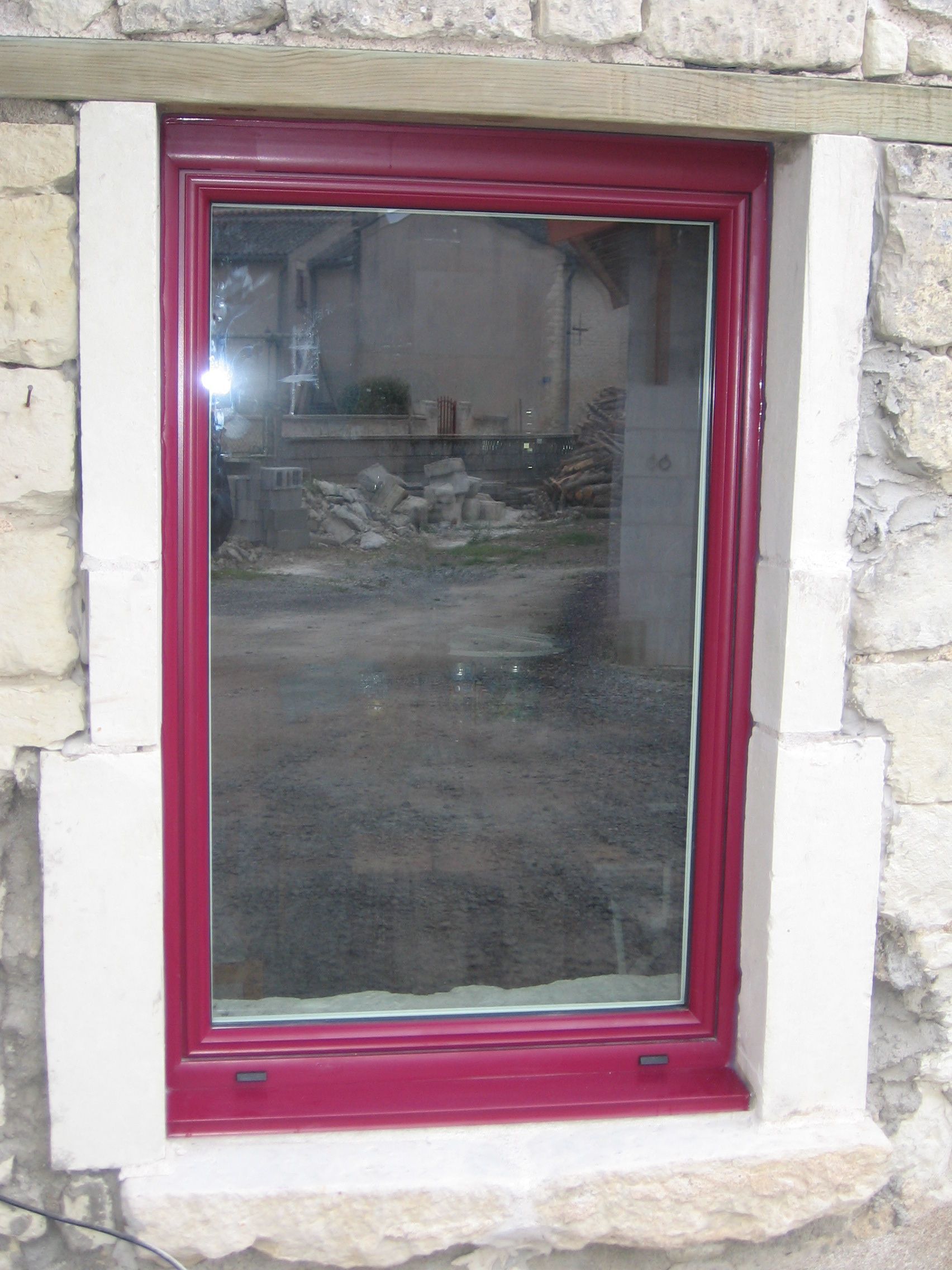 Une fenêtre rectangulaire verticale à cadre magenta foncé, encastrée dans un mur de pierre rustique de couleur claire.