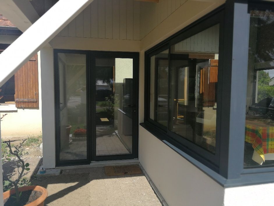 Vue extérieure de l'entrée d'une maison avec une porte vitrée à cadre noir et un coin de fenêtre sur un mur clair.