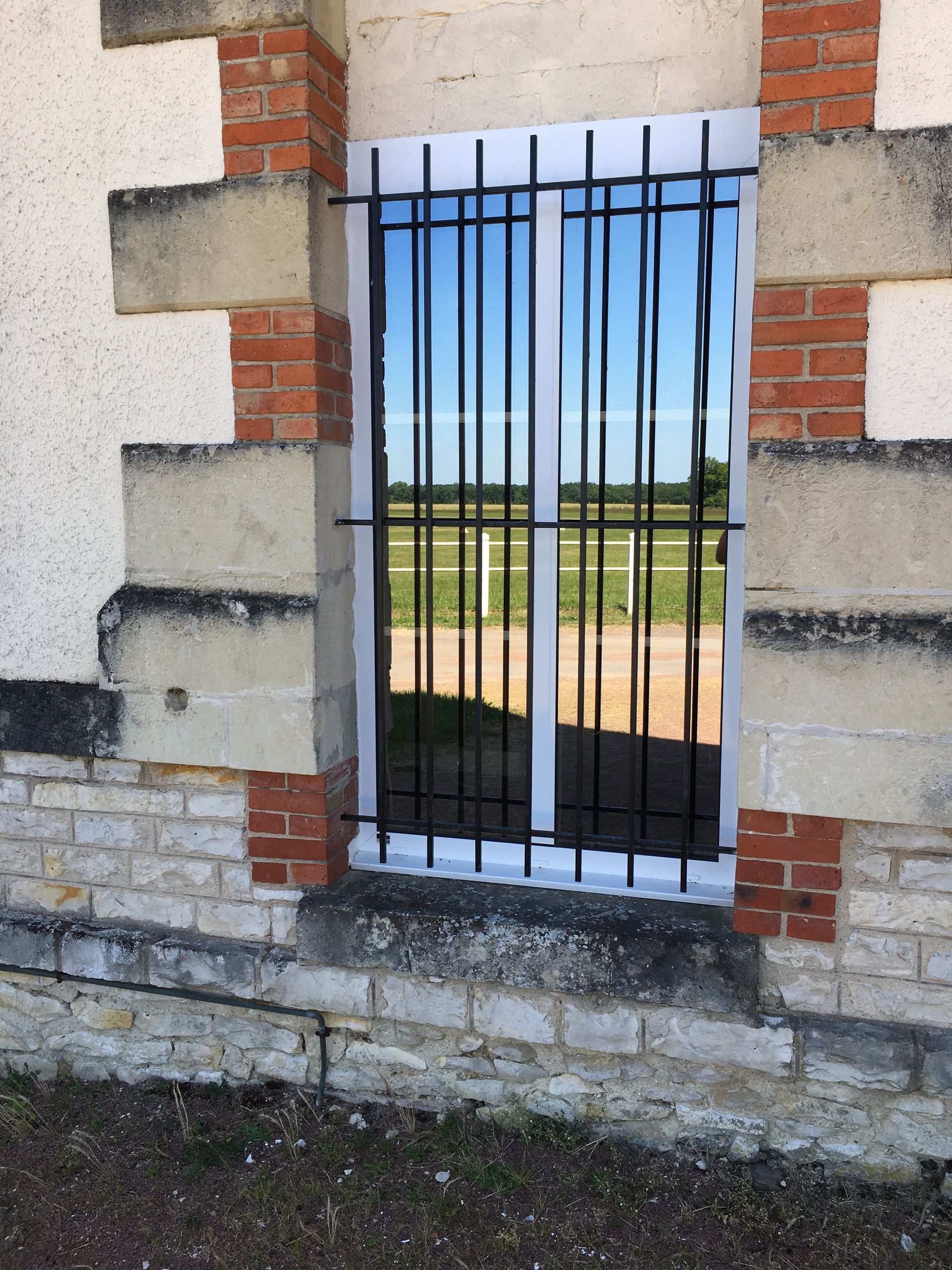 Une fenêtre rectangulaire avec une grille de sécurité en fer noir, encastrée dans un mur de pierre et de brique donnant sur un champ herbeux.