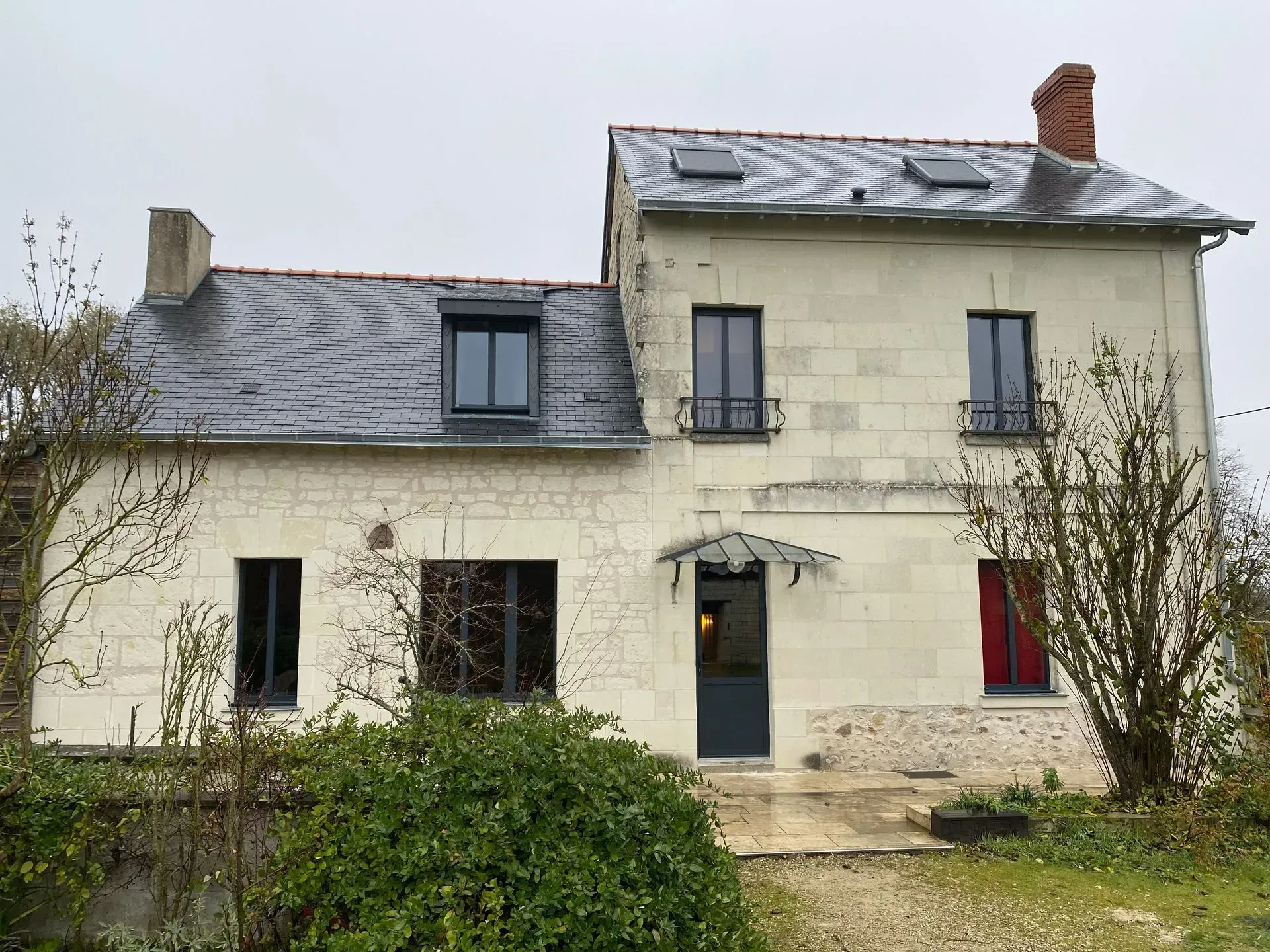 Maison en pierre de deux étages avec un toit en ardoise, des volets gris et une entrée avec un petit auvent métallique donnant sur un chemin de gravier.