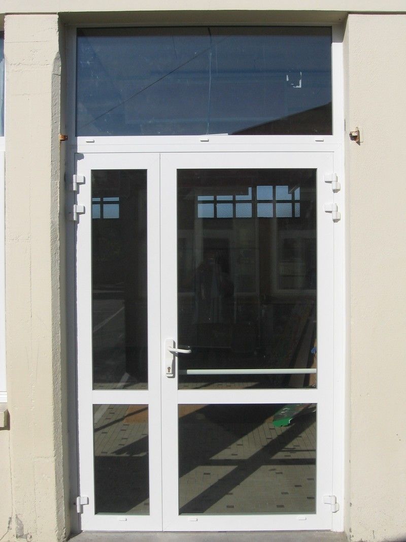 Une porte vitrée à cadre blanc, avec un panneau latéral et une imposte rectangulaire au-dessus, encastrée dans un mur clair et uni.