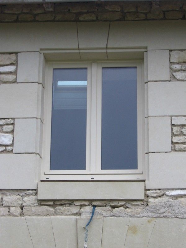 Une fenêtre rectangulaire à l'encadrement beige clair, encastrée dans un mur de maçonnerie de pierres mixtes et de grands blocs de pierre lisses.