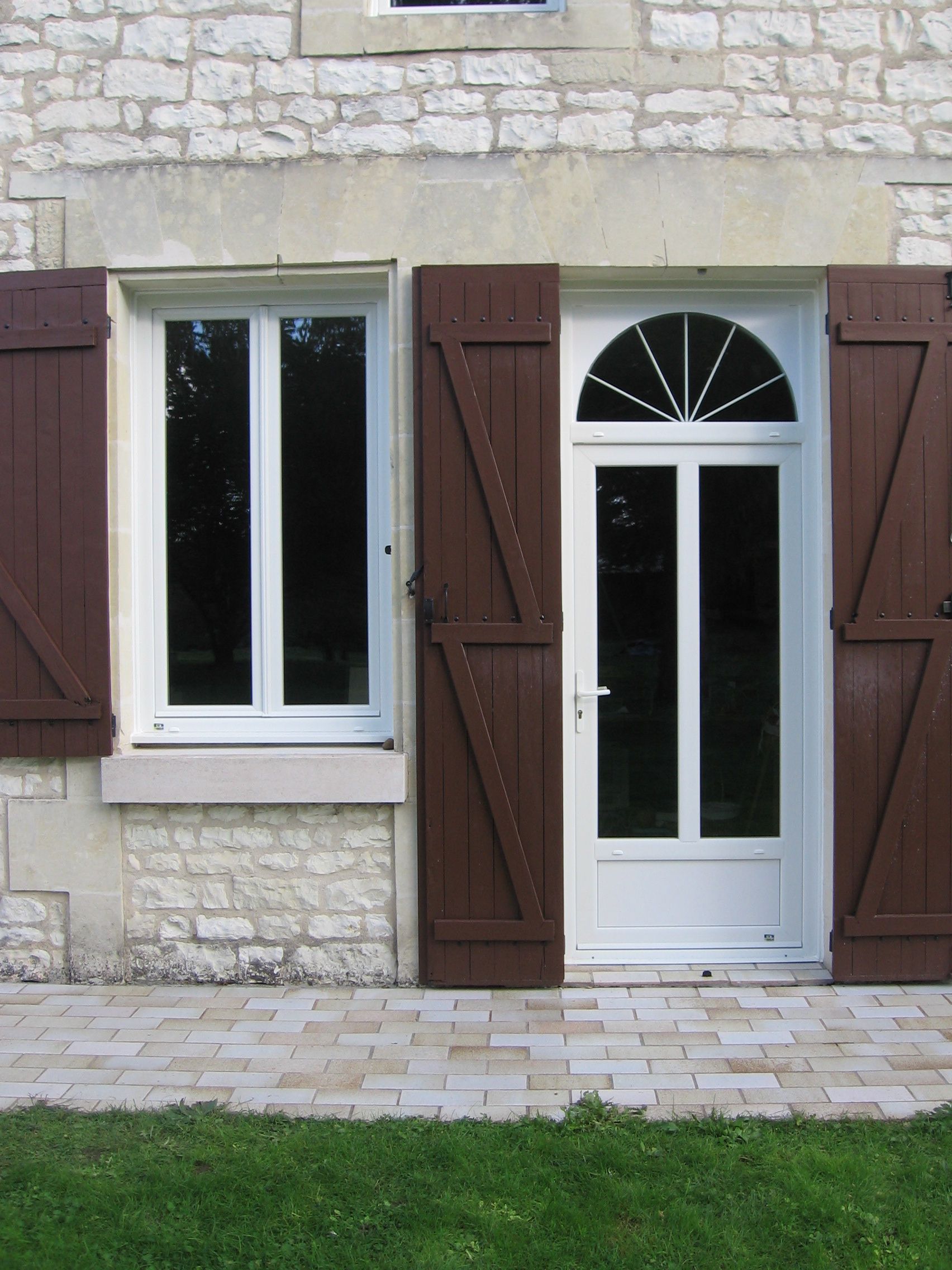L'extérieur d'une maison en pierre présente une porte blanche ornée d'une imposte et d'une fenêtre simple, toutes deux encadrées de volets bruns.
