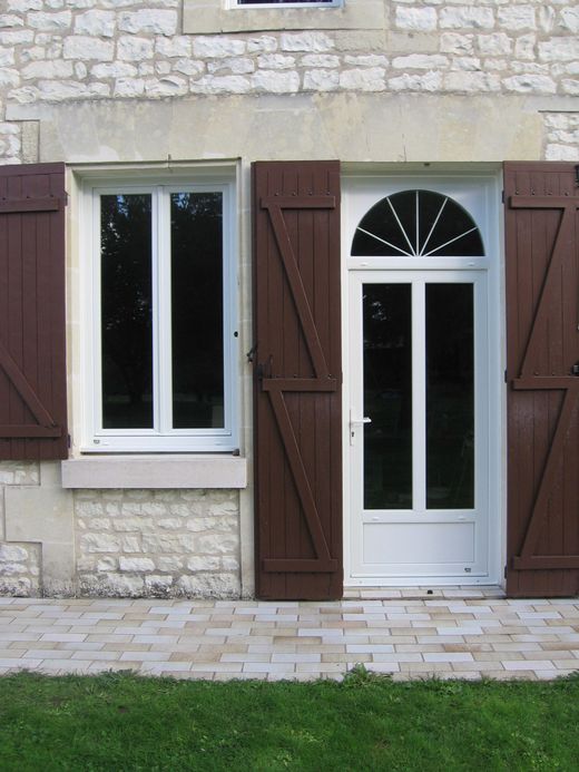 L'extérieur d'une maison en pierre blanche présente une fenêtre rectangulaire et une porte surmontée d'une imposte, toutes deux dotées de volets en bois brun.