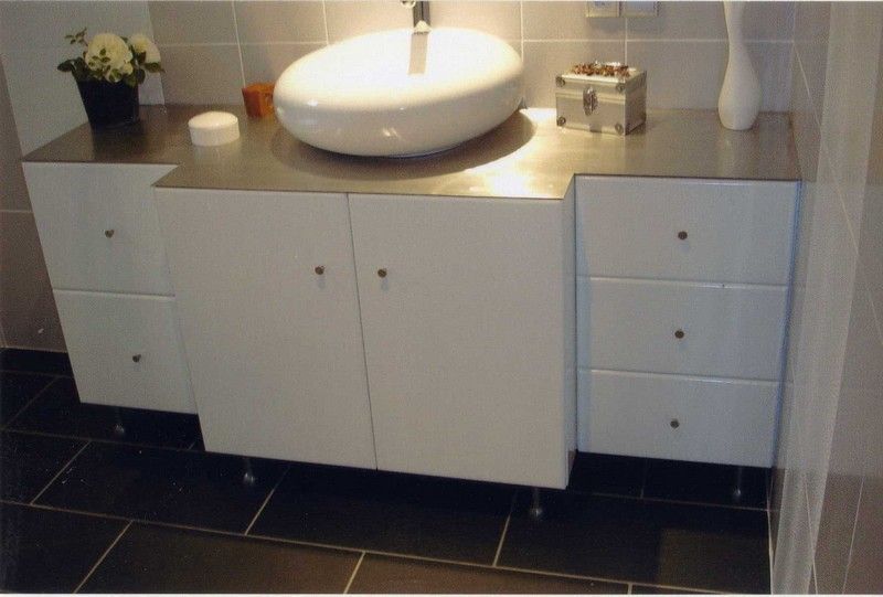 Un meuble-lavabo moderne avec une vasque blanche sur un plan de travail en métal, flanqué de tiroirs et d'armoires blanches.