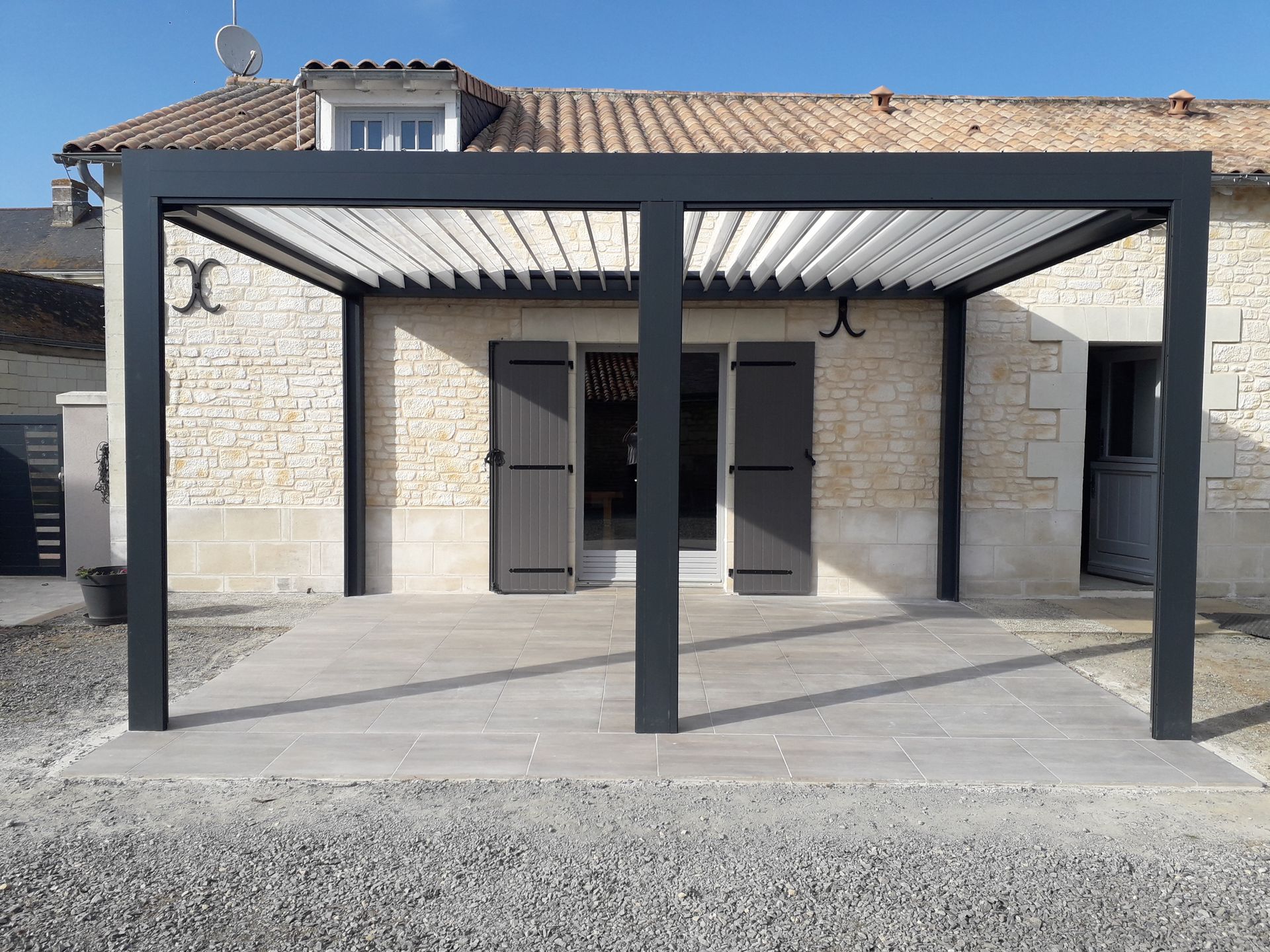 Une pergola noire moderne, dotée d'un toit à lames orientables rétractables, surplombe une terrasse carrelée devant une maison en pierre.