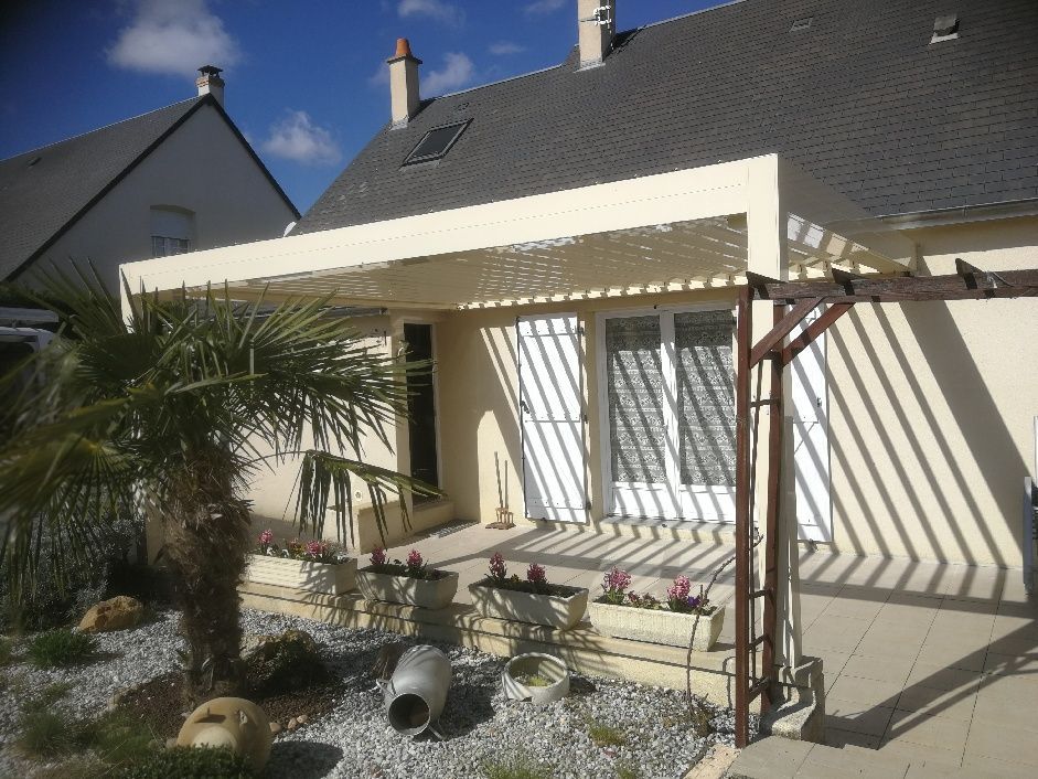 Une maison beige avec un patio, une pergola à claire-voie, un palmier et un petit parterre de fleurs par une journée ensoleillée.