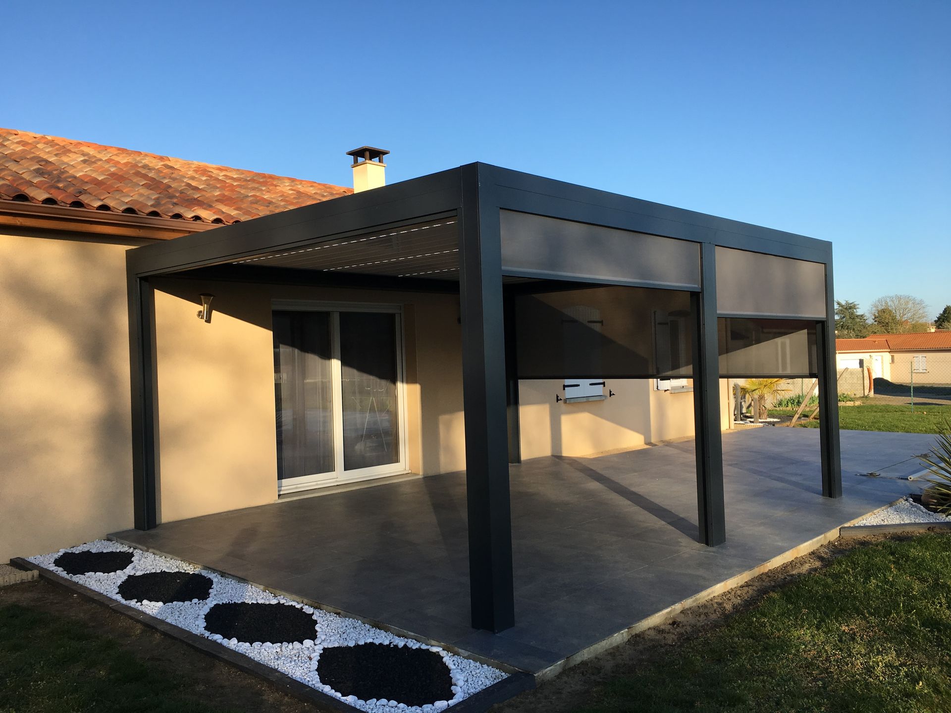 Une pergola de patio à cadre sombre avec des écrans latéraux rétractables, fixée au mur extérieur d'une maison au toit de tuiles.