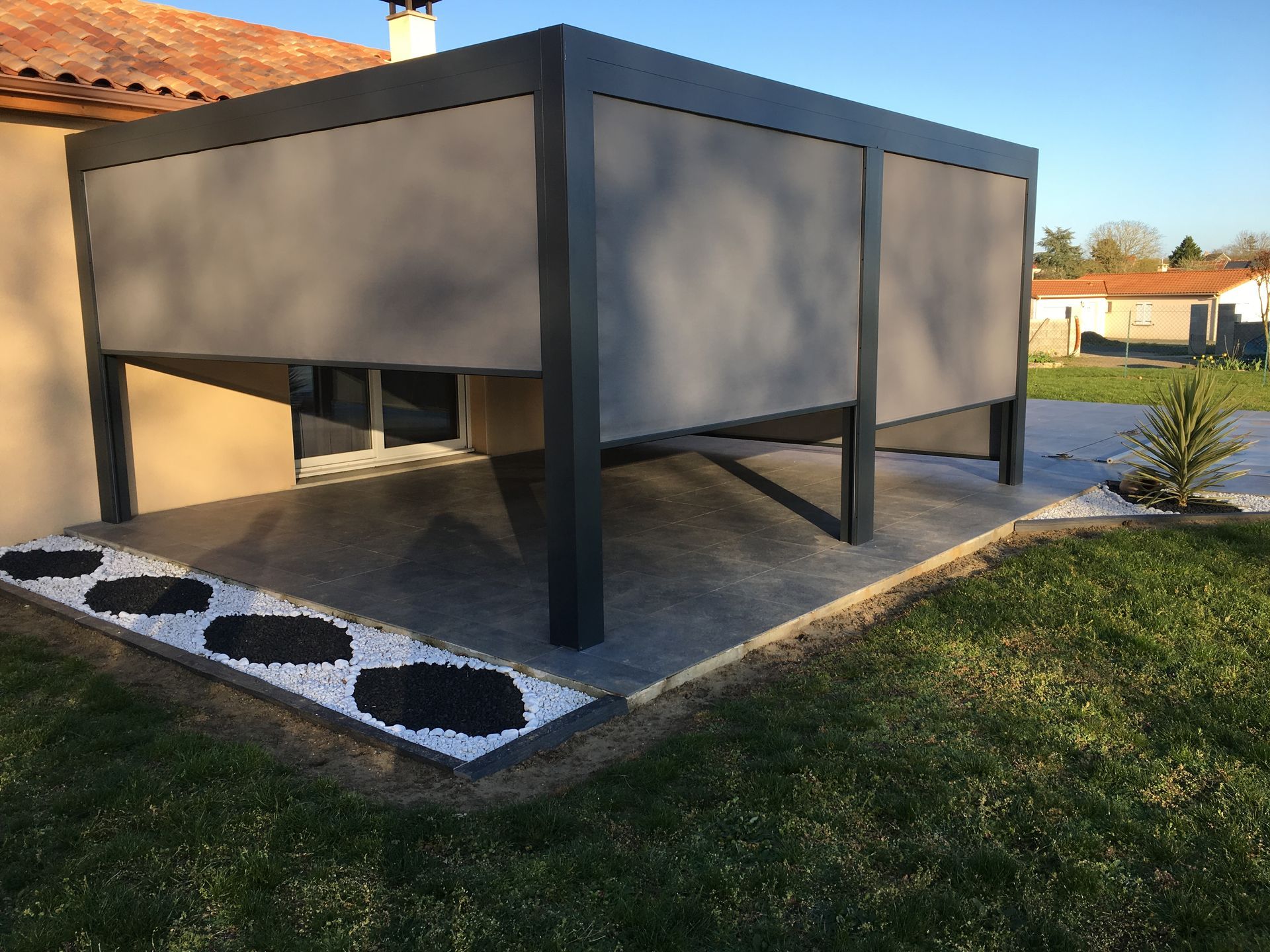 Une pergola noire moderne avec des panneaux d'intimité gris sur une terrasse en pierre à côté d'une maison, entourée de pelouse.