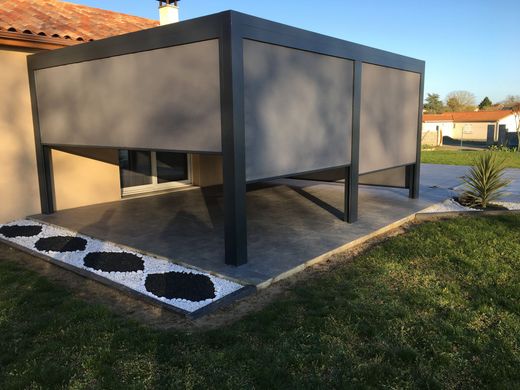 Une pergola gris foncé avec des auvents latéraux prolongés est installée sur une terrasse carrelée à côté d'une maison, entourée d'une pelouse et de gravier blanc.