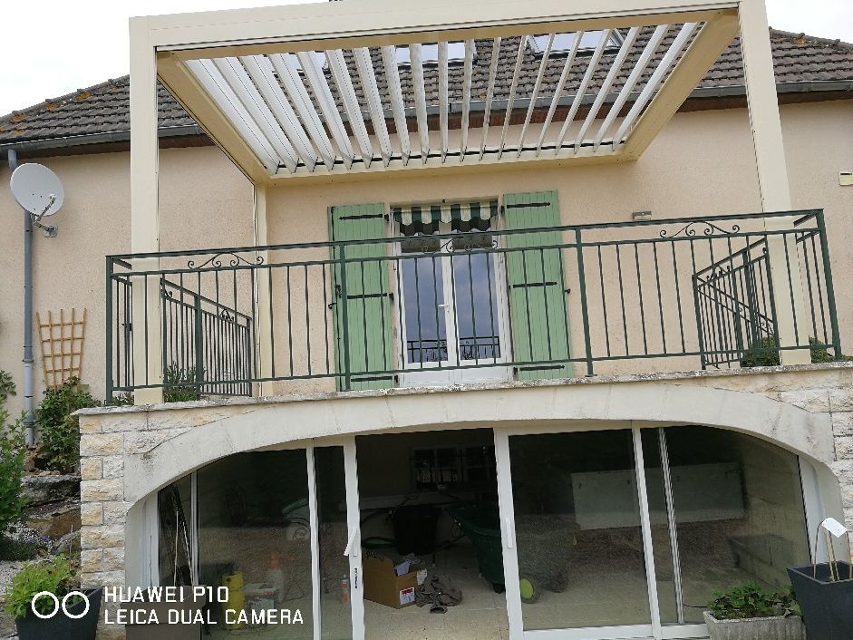 L'extérieur d'une maison avec un patio ouvert en contrebas, un balcon supérieur à balustrade agrémenté d'une pergola à persiennes et de volets verts.