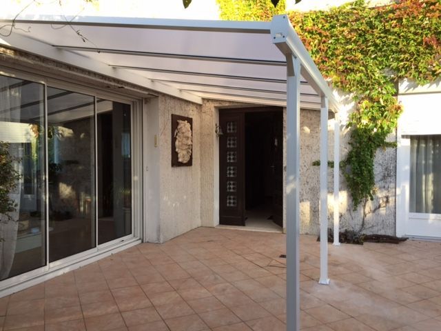 Un patio avec un auvent blanc, incliné et translucide, recouvrant une zone carrelée à côté d'une porte vitrée et d'un mur recouvert de lierre.