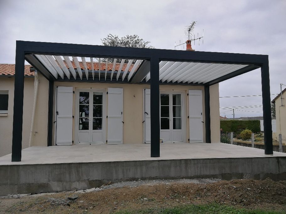 Une pergola noire à persiennes installée sur une terrasse en béton devant une maison aux portes doubles blanches.