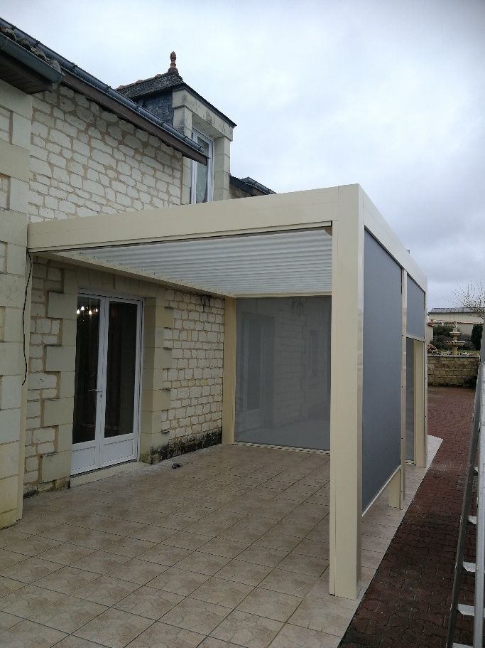Une pergola beige à persiennes, adossée à une maison en pierre, avec des panneaux latéraux pour préserver l'intimité et une terrasse pavée.