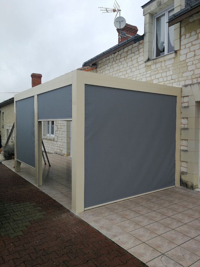 Une pergola de couleur beige, adossée à une maison en pierre, avec des écrans verticaux gris partiellement déployés pour préserver l'intimité.