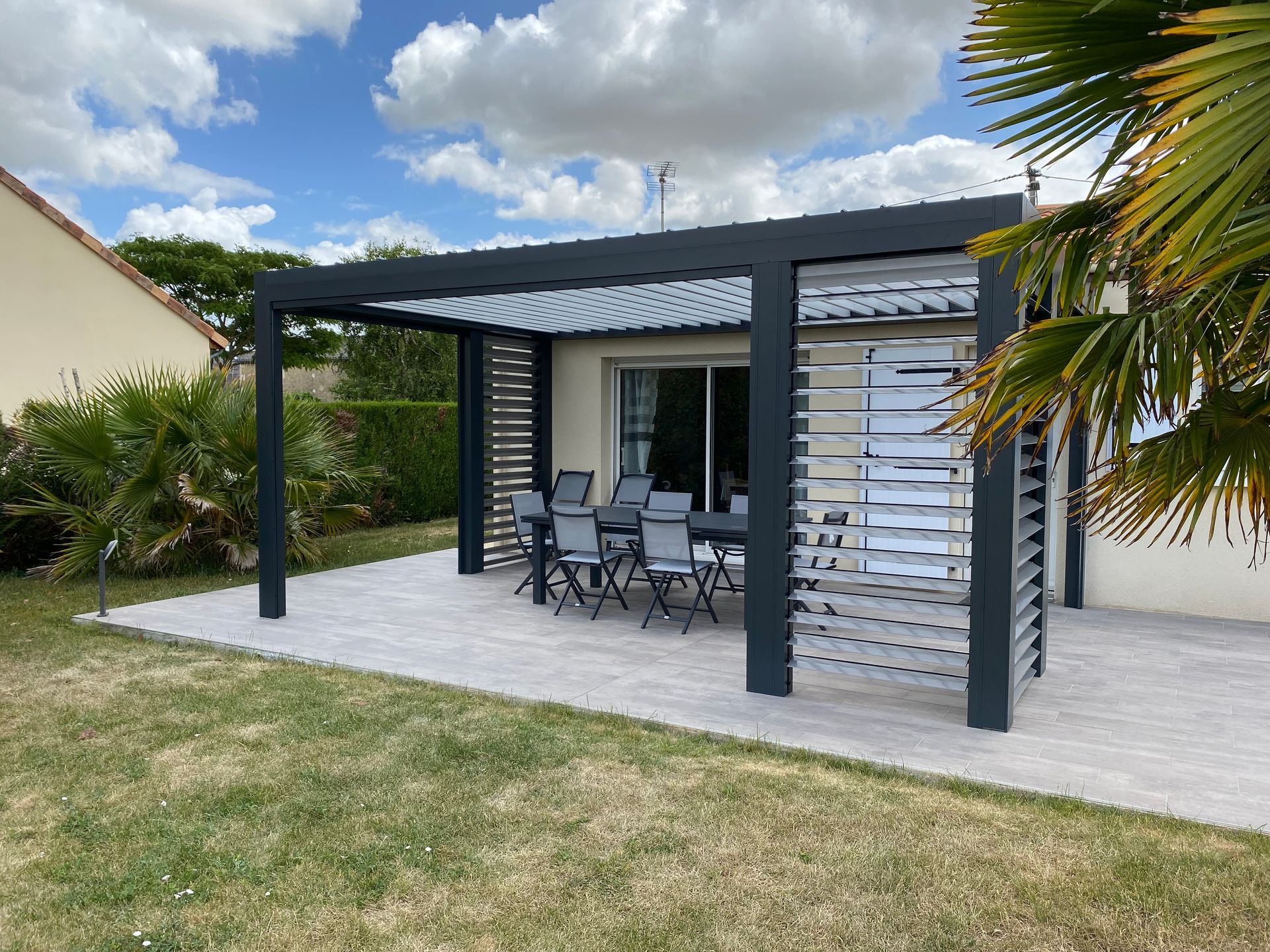 Une pergola moderne noire à persiennes avec un ensemble de salle à manger sur une terrasse en pierre dans un jardin gazonné.