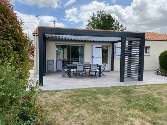 Un patio agrémenté d'une pergola noire moderne abritant une table et des chaises de salle à manger, le tout adossé à la façade d'une maison.