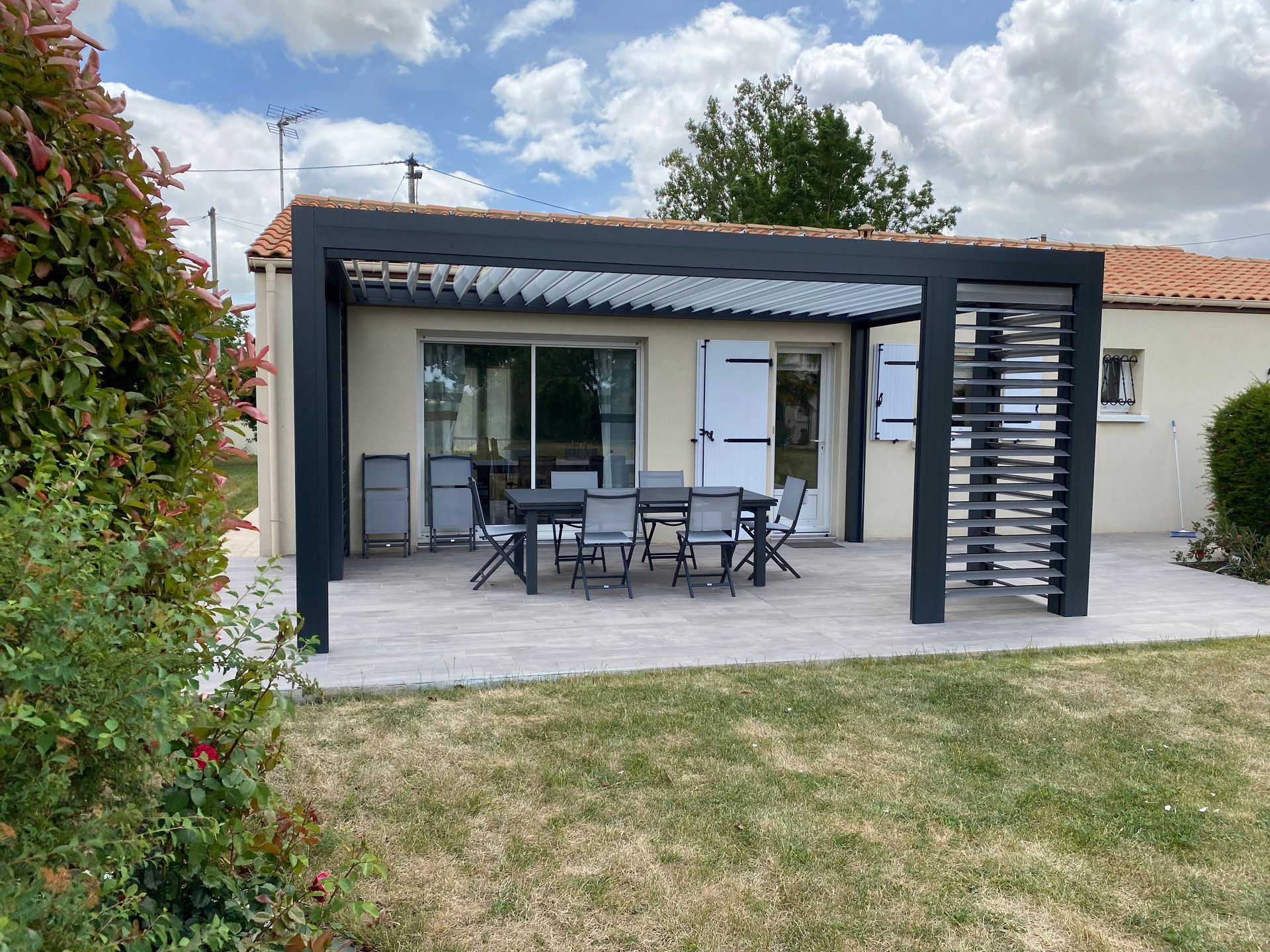 Une terrasse avec une pergola noire moderne, un ensemble de salle à manger et un paravent, attenante à une maison aux volets blancs.