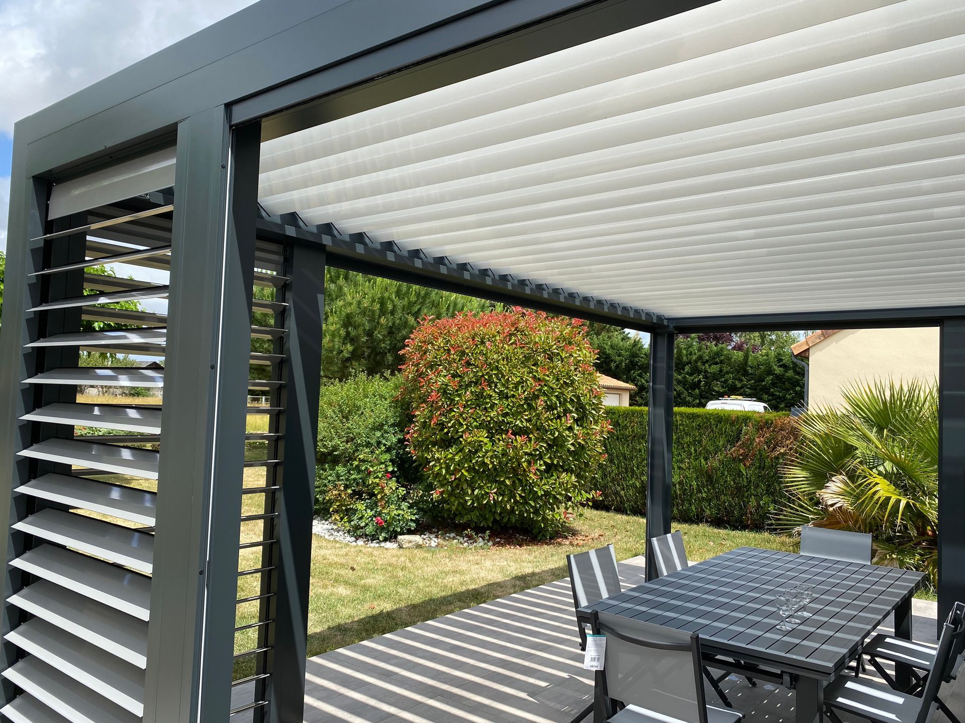 Une pergola moderne gris foncé avec toit et panneaux latéraux à lames orientables, au-dessus d'un ensemble de patio dans un jardin.