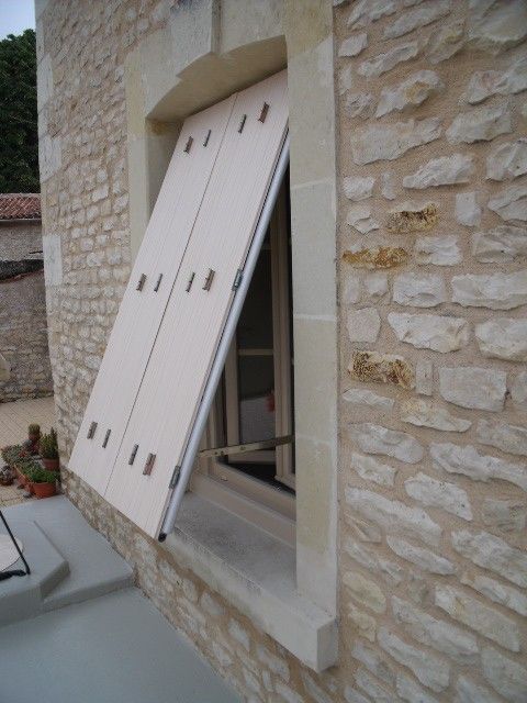 Un volet en bois clair est maintenu ouvert sur un bâtiment en pierre, révélant une ouverture de fenêtre.