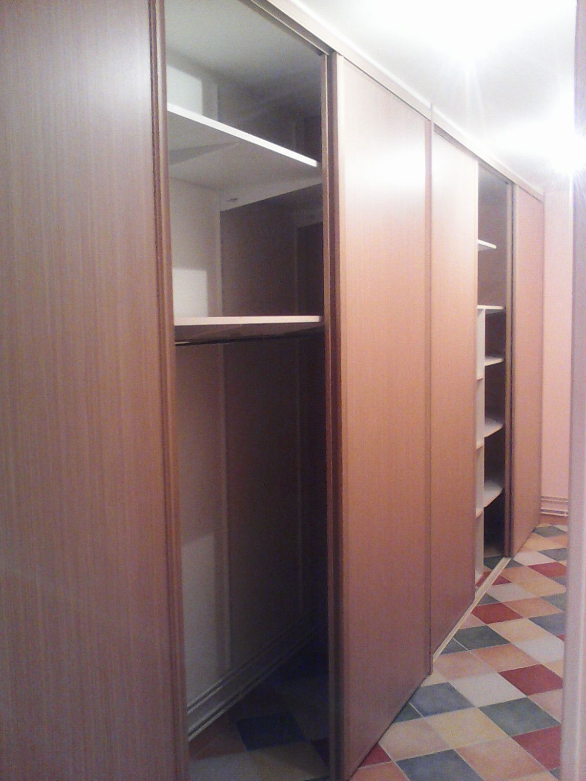 Une grande armoire en bois clair veiné, avec des portes coulissantes ouvrantes et fermantes, placée dans un couloir au sol carrelé à damier.