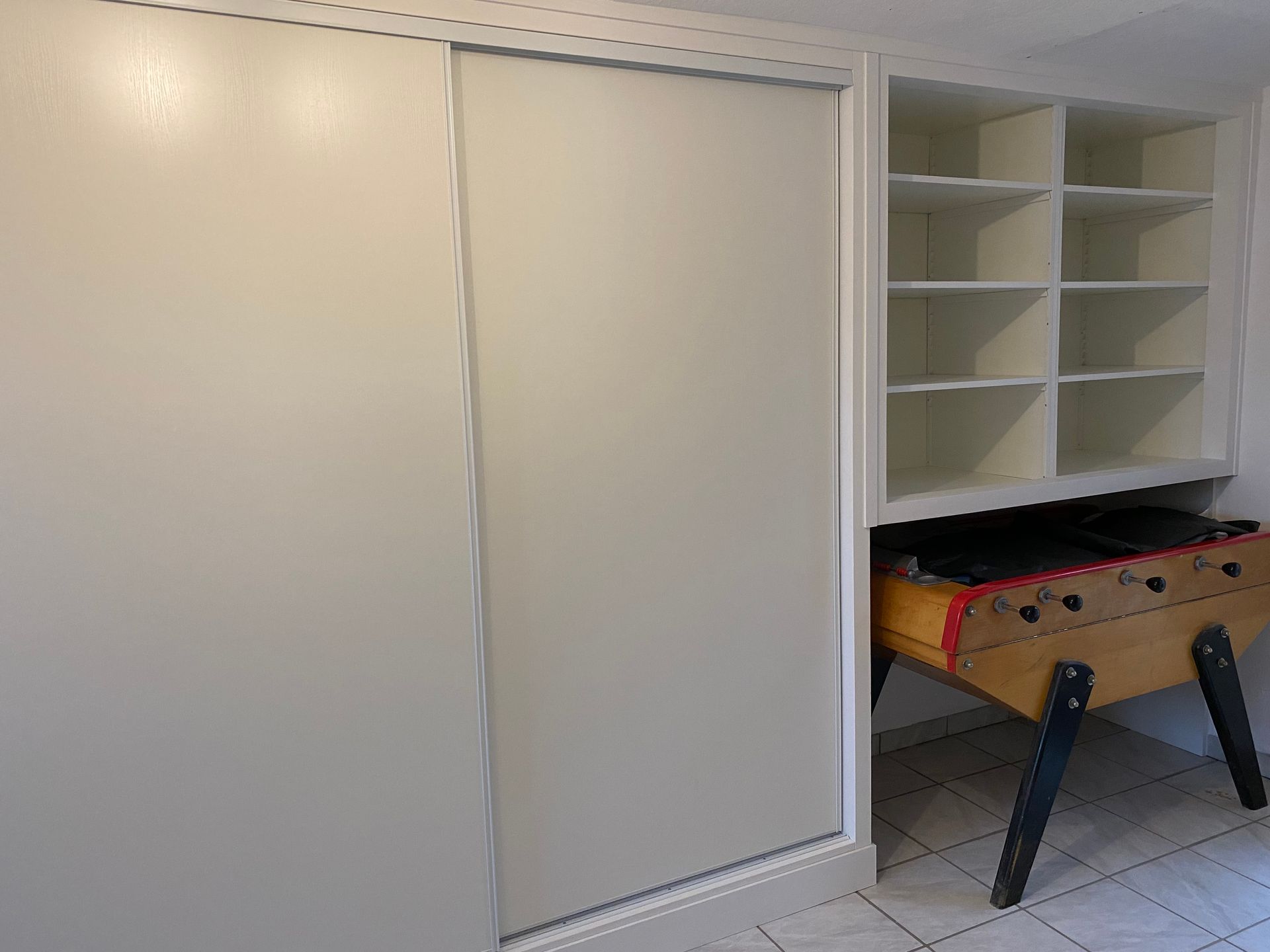 Des portes de placard coulissantes blanches jouxtent une étagère blanche au-dessus d'un baby-foot ; un sol carrelé.