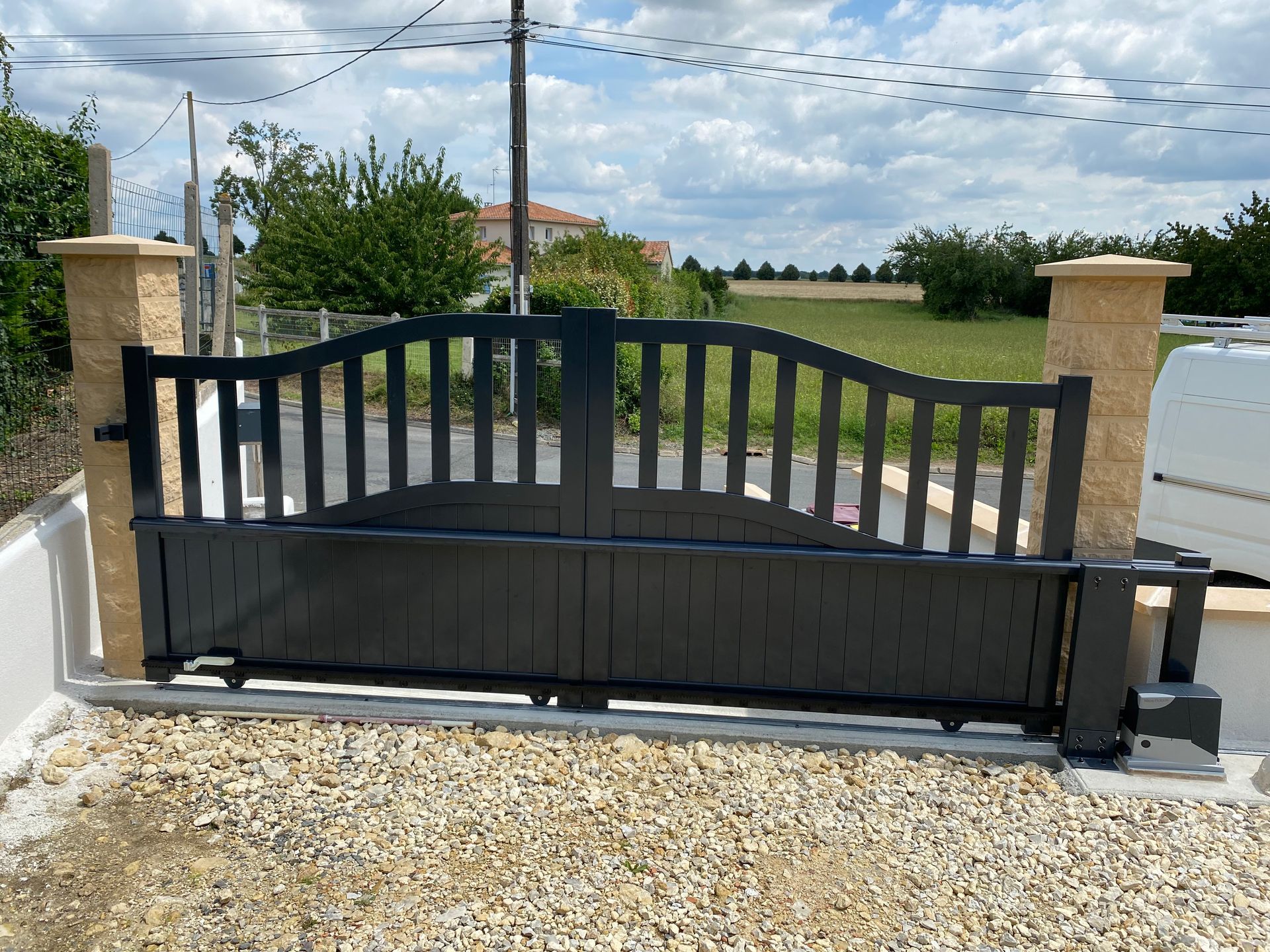 Un portail coulissant en métal noir à lattes verticales se dresse entre deux piliers de pierre sur une allée de gravier, sous un ciel nuageux.