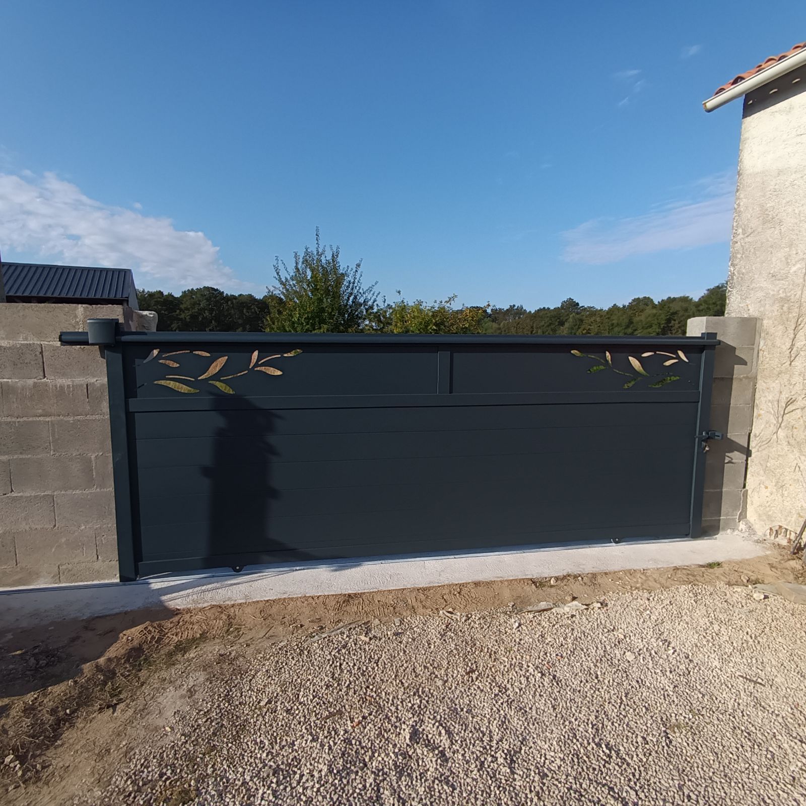 Un portail moderne gris foncé orné de motifs à la feuille d'or, installé entre des piliers en béton sur une allée de gravier.